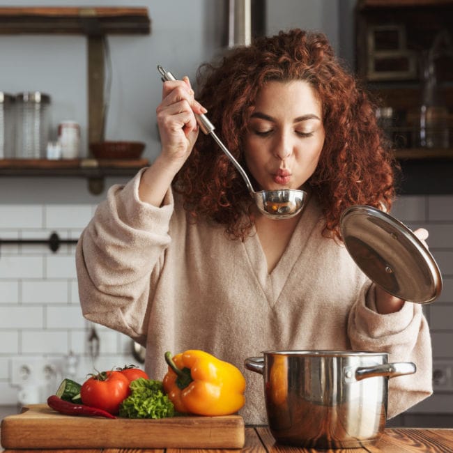 woman tasting soup in kitchen with vegetables on the island