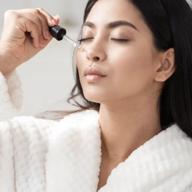 woman applying hyaluronic acid serum to face with hands white robe dark hair
