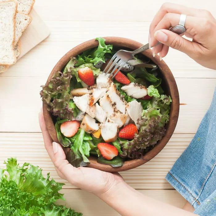 woman sitting on floor eating salad topped with chicken and strawberries