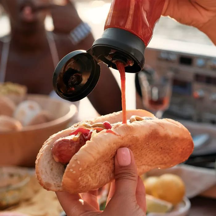 Pouring ketchup onto a hot dog on a picnic table.