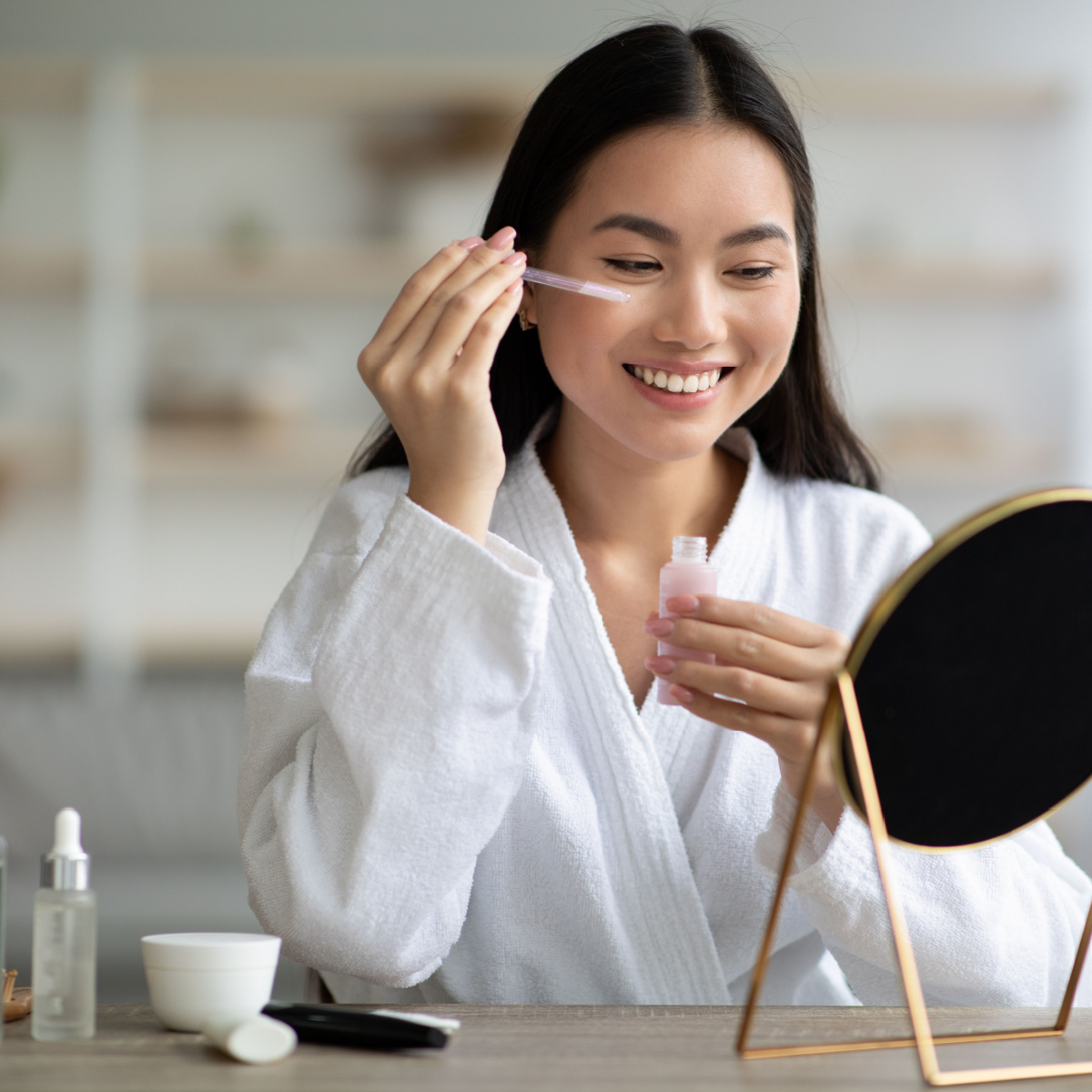 woman applying serum to her dark spots under-eyes white robe long brown hair mirror stand-up table cosmetics