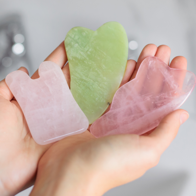 hand holding pink and green gua sha tools over sink faucet