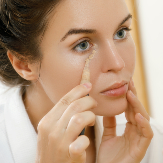 woman adding thin layer of concealer to her under-eyes looking in mirror