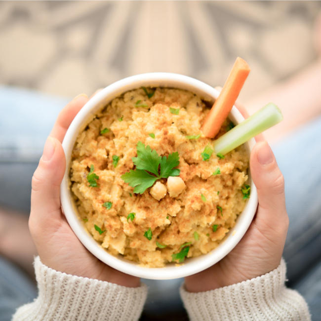 hands holding white bowl of tan hummus chickpeas parsley carrots celery dip