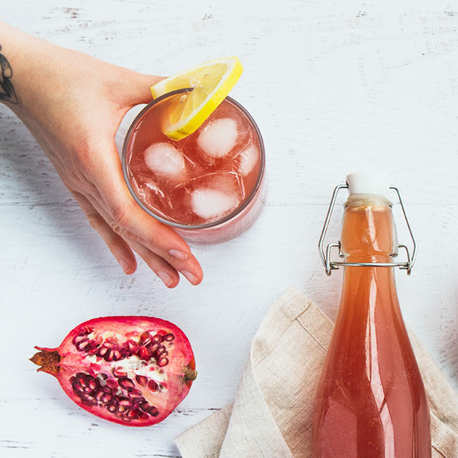 pomegranate kombucha bottle poured into glass with ice cubes lemon chopped pomegranate on white table hand holding glass