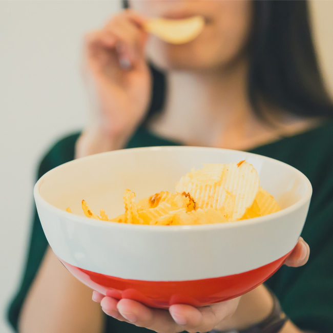 woman holding out red and white bowl of yellow chips and holding chip in hand biting into chip brown hair green shirt