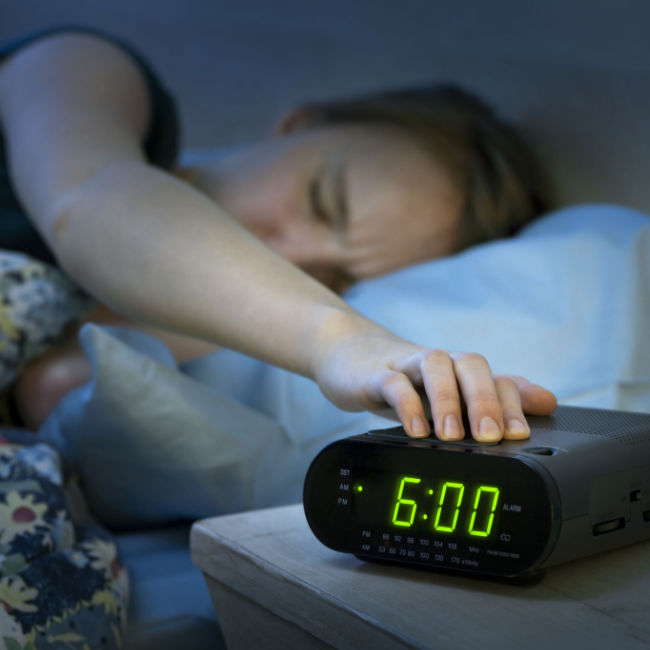 woman in bed hitting snooze on digit alarm clock at 6 AM