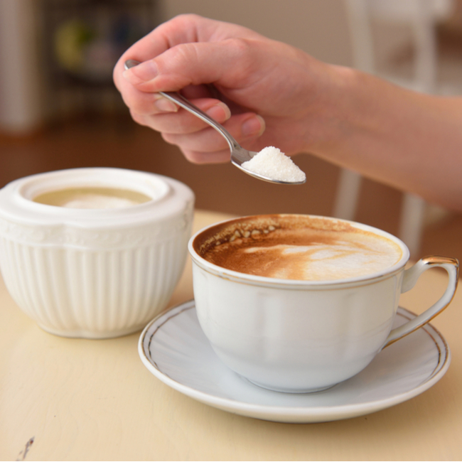 person putting spoonful of sugar into mug of coffee