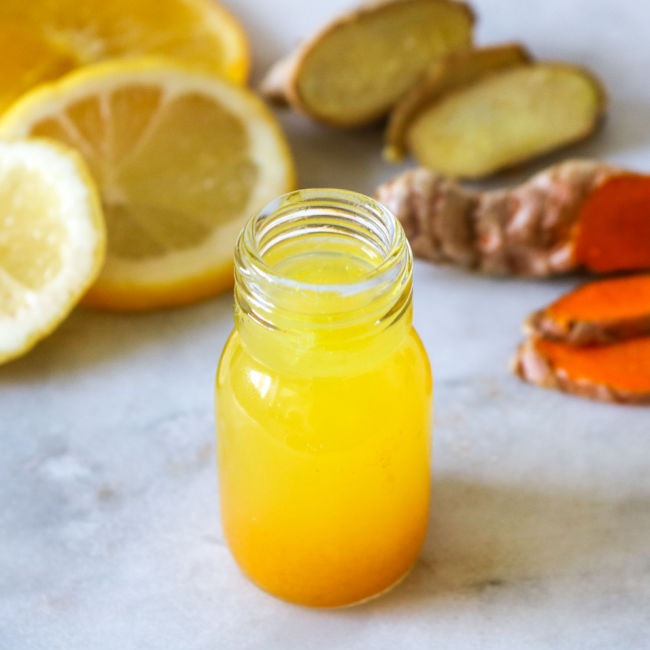 small glass bottle of orange/yellow drink with ginger and lemon behind it