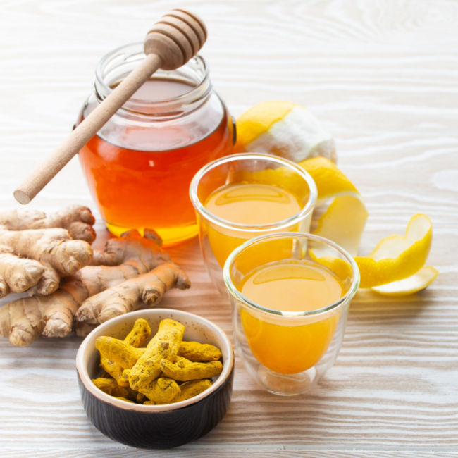 two shot glasses filled with orange/yellow drink surrounded by ginger, honey, and lemon