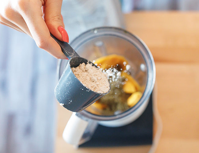 hand pouring scoop of protein powder into smoothie