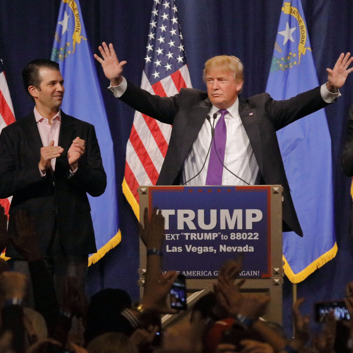 donald trump waving hands in air while speaking blue tuxedo red tie american flag podium