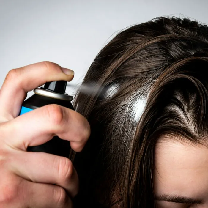 woman spraying white dry shampoo directly onto brown greasy oily hair