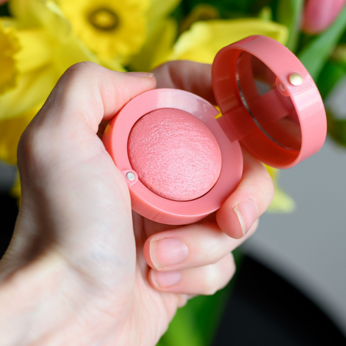 hand holding compact circular pink blush with flowers in background