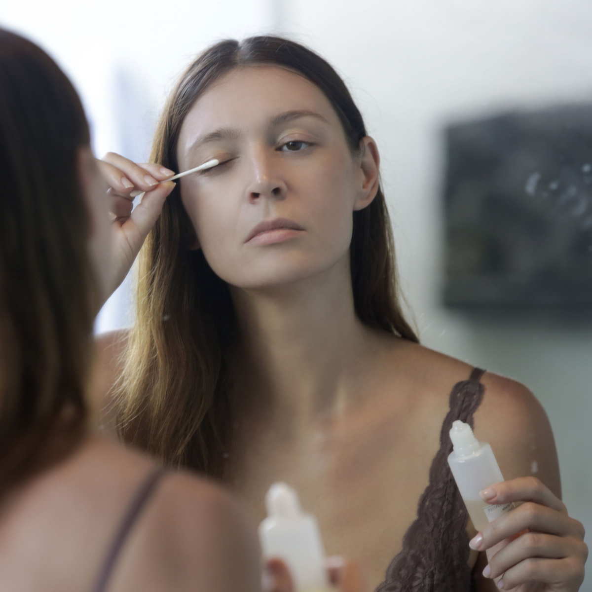 woman applying eyelash serum mirror brown hair brown tank top cosmetic product