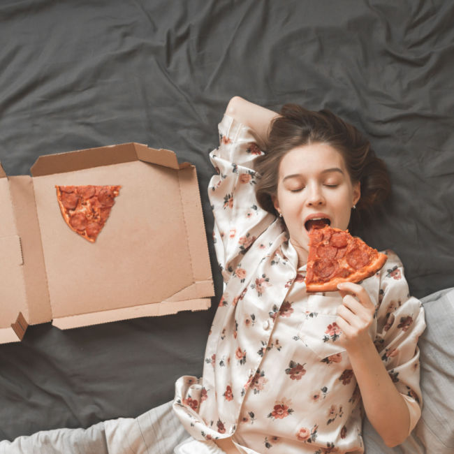 woman eating pizza out of box in bed