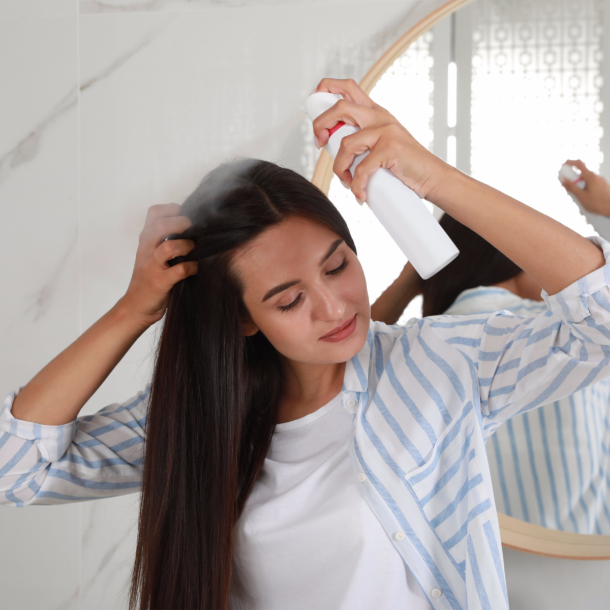 woman spraying dry shampoo onto scalp long brown hair