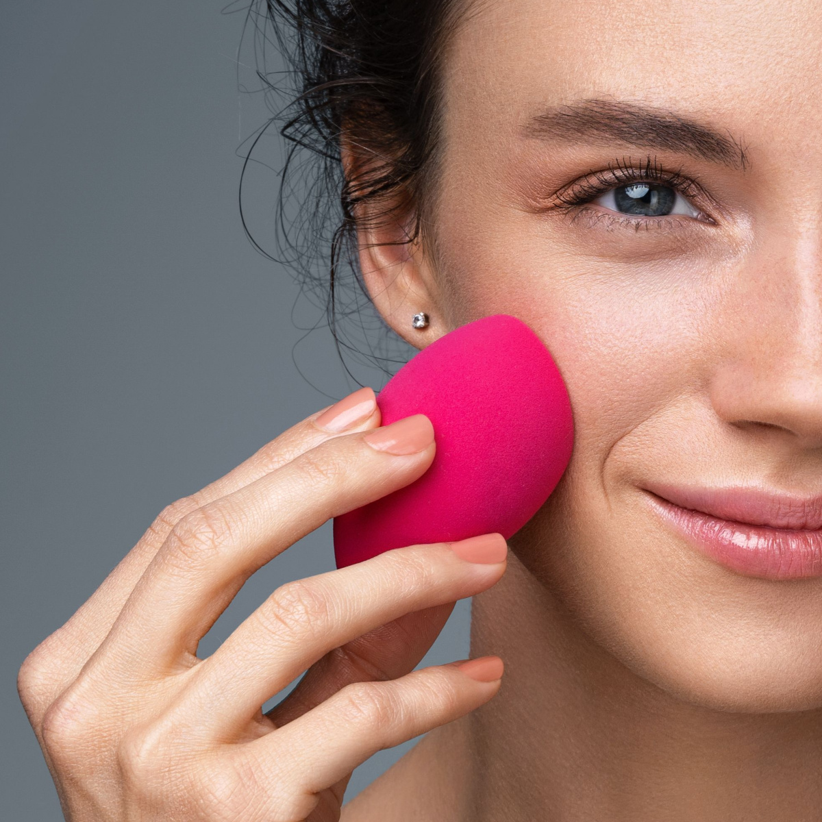 woman using bright pink beauty blender makeup sponge applying product blending cheekbone gray background brown hair blue eyes
