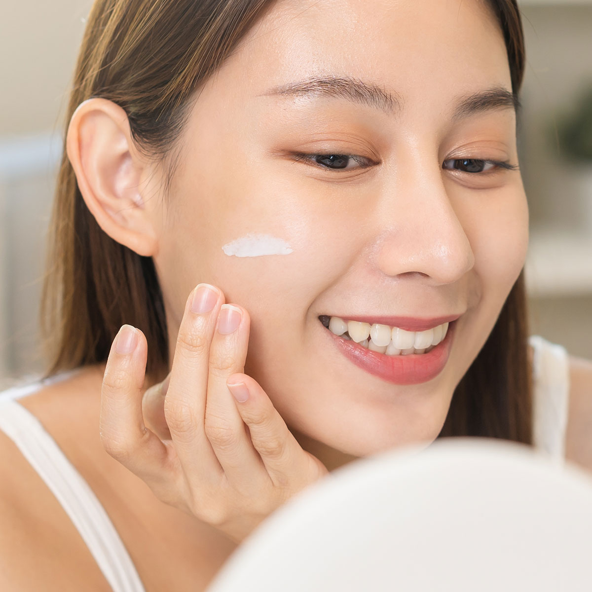 young woman applying white facial moisturizer sunscreen primer product in front of mirror