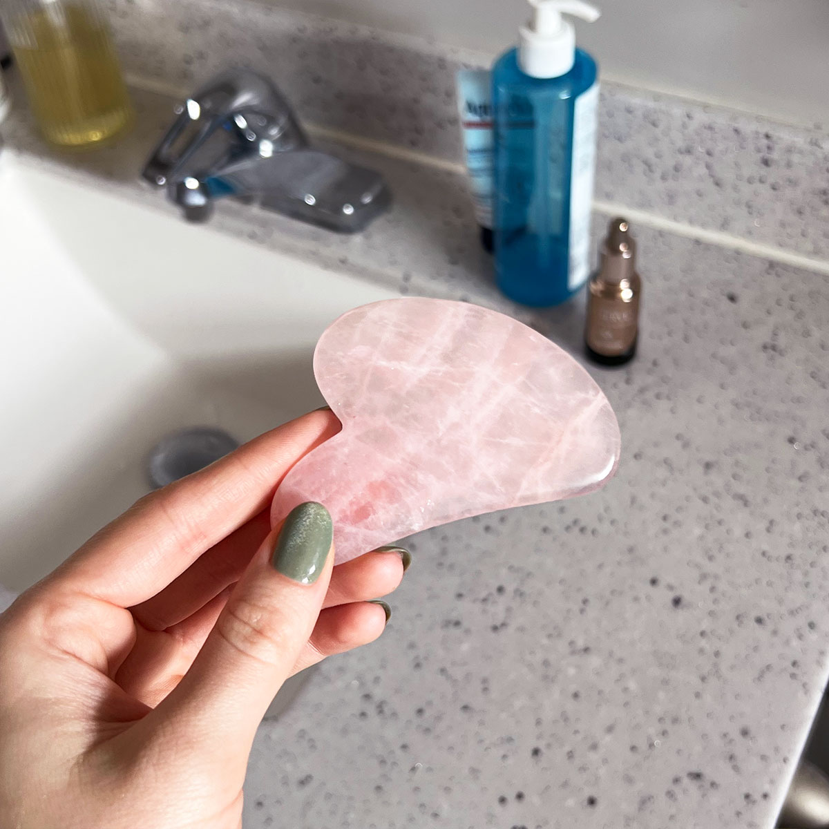 manicured hand holding pink gua sha sculpting tool over bathroom sink