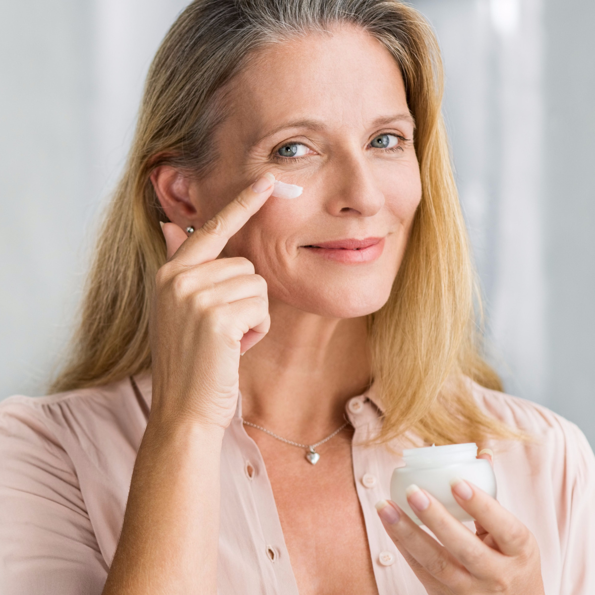 senior woman with long hair applying eye cream with finger to under-eye skin from small container