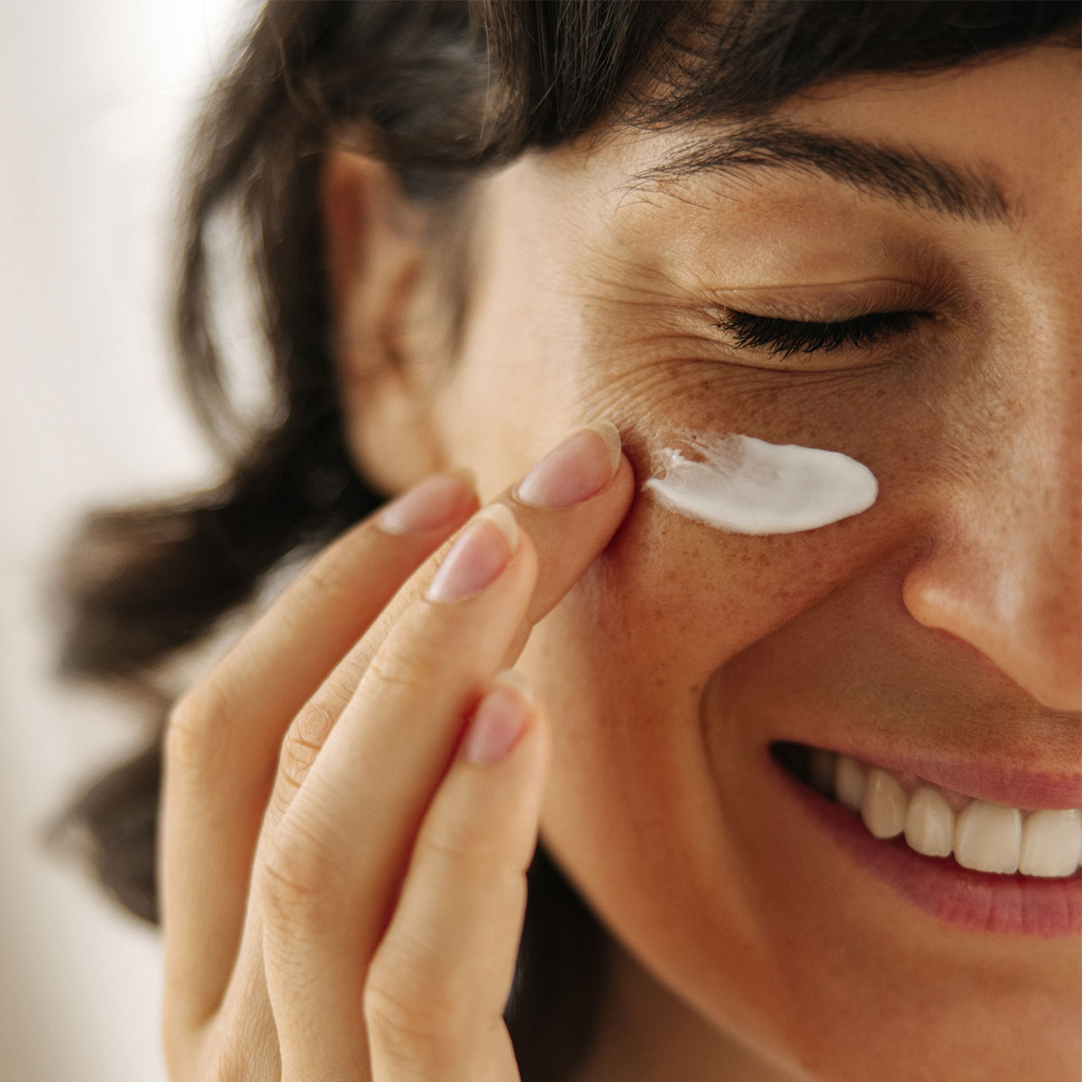woman smiling and applying white under eye cream to skin with finger
