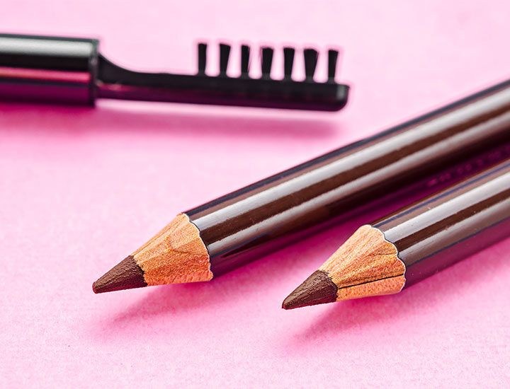 two brown eyebrow pencils and eyebrow brush on pink background isolated