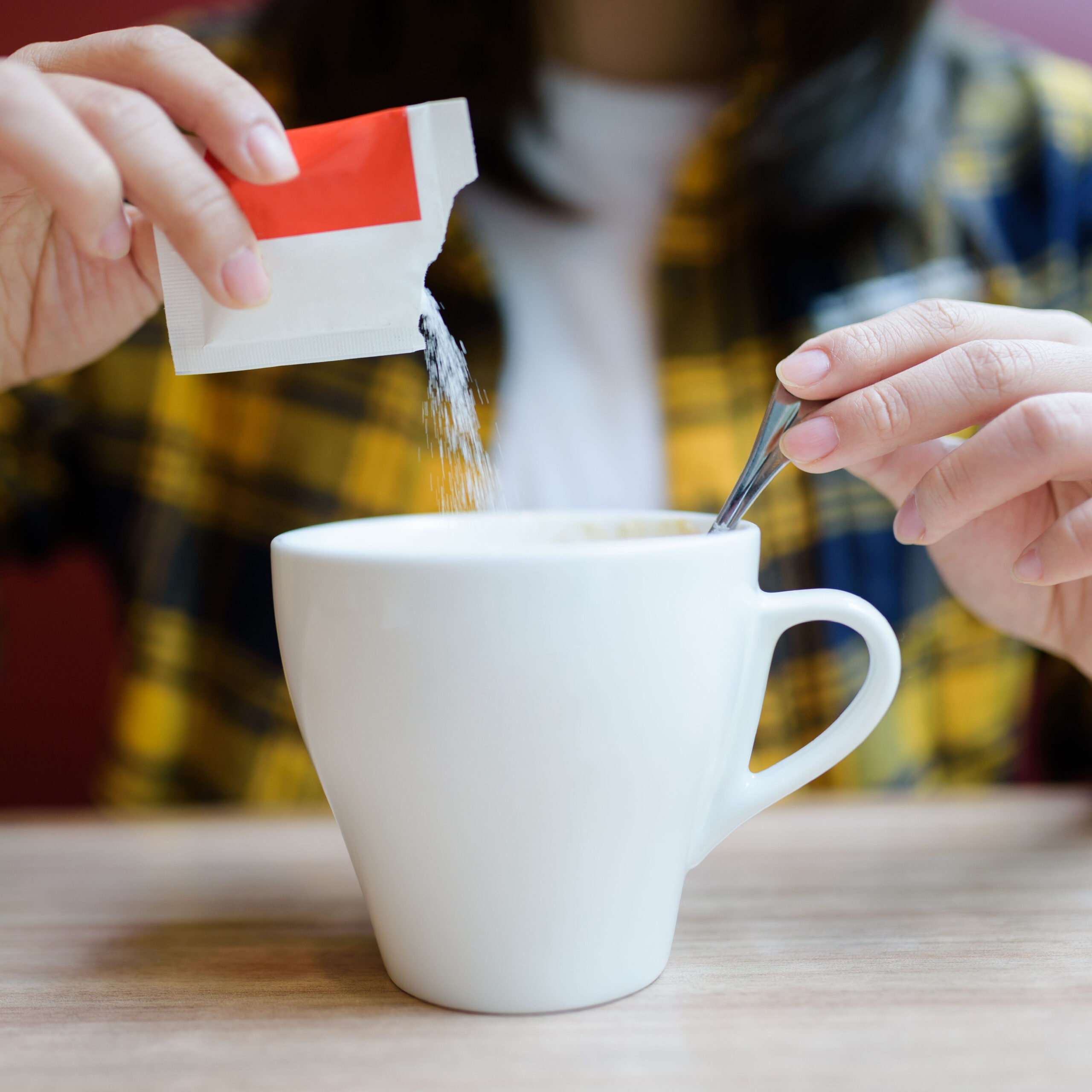 person pouring artificial sweetener packet into cup of coffee