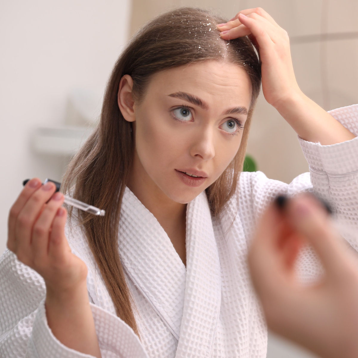 woman applying scalp oil hand on head white robe looking in mirror