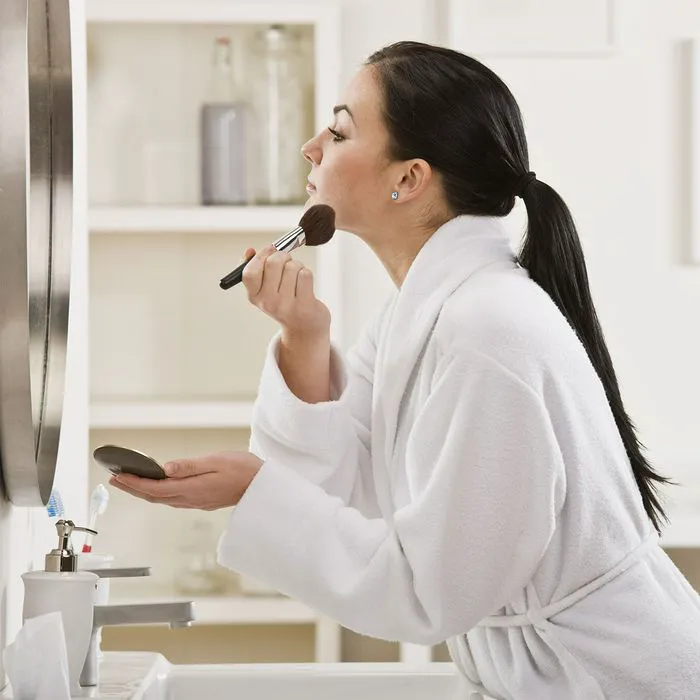 "Woman putting on makeup in front of a mirror while wearing a bathrobe"