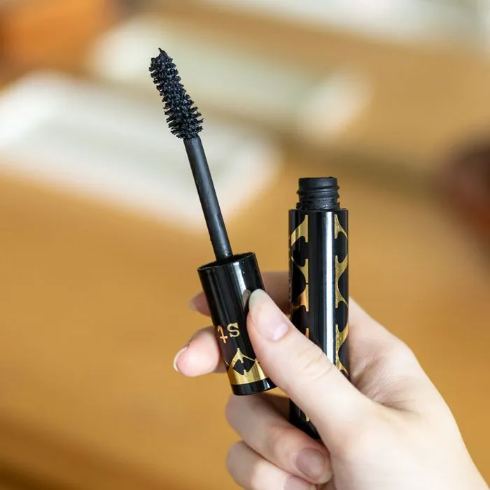 Hand holding a mascara tube and wand against a neutral background.
