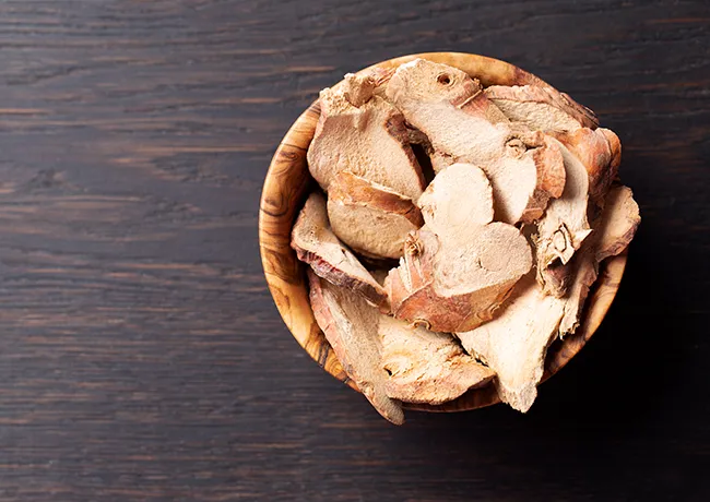 Dried galangal slices in a wooden bowl on dark table.
